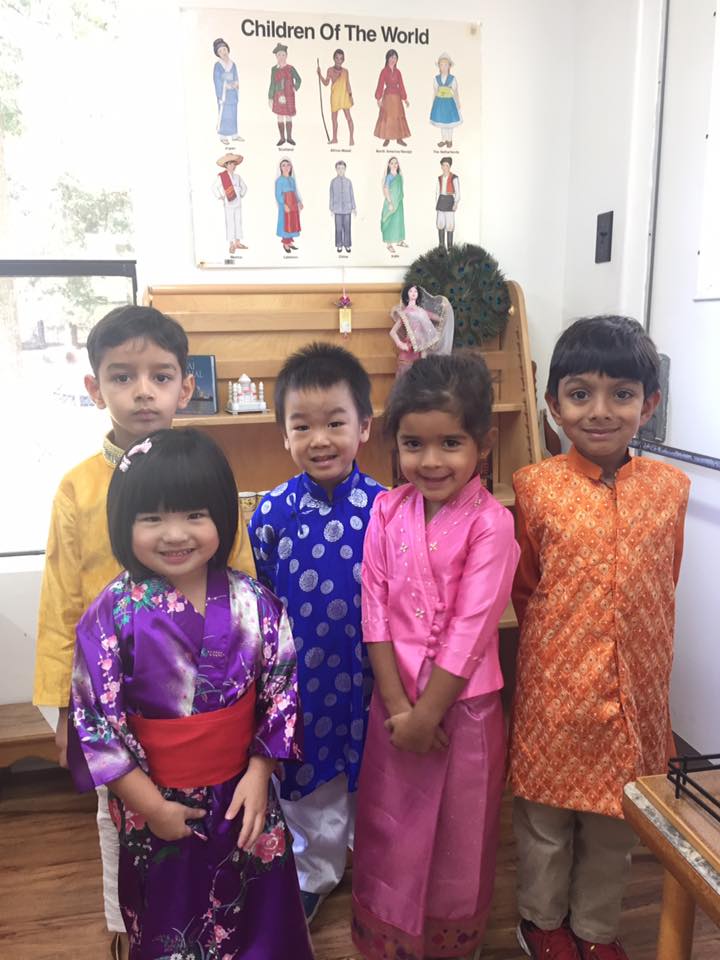 Montessori Children in diverse cultural attire pose in a classroom, with a world cultures poster in the background.