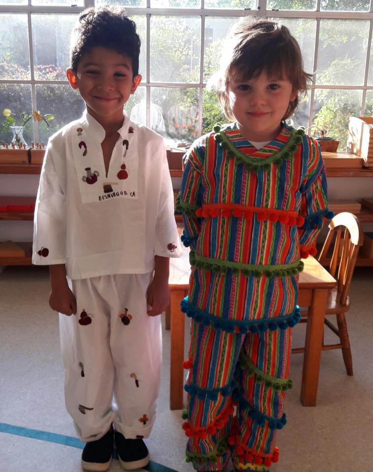 Two Montessori children in costumes stand near a window, smiling. The costumes are white with small black details and striped with pom poms.