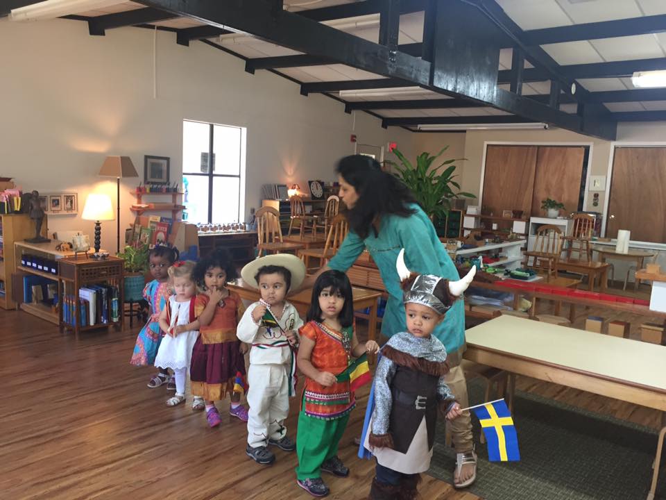 Montessori Children in costumes line up with a teacher, holding flags, in a classroom.