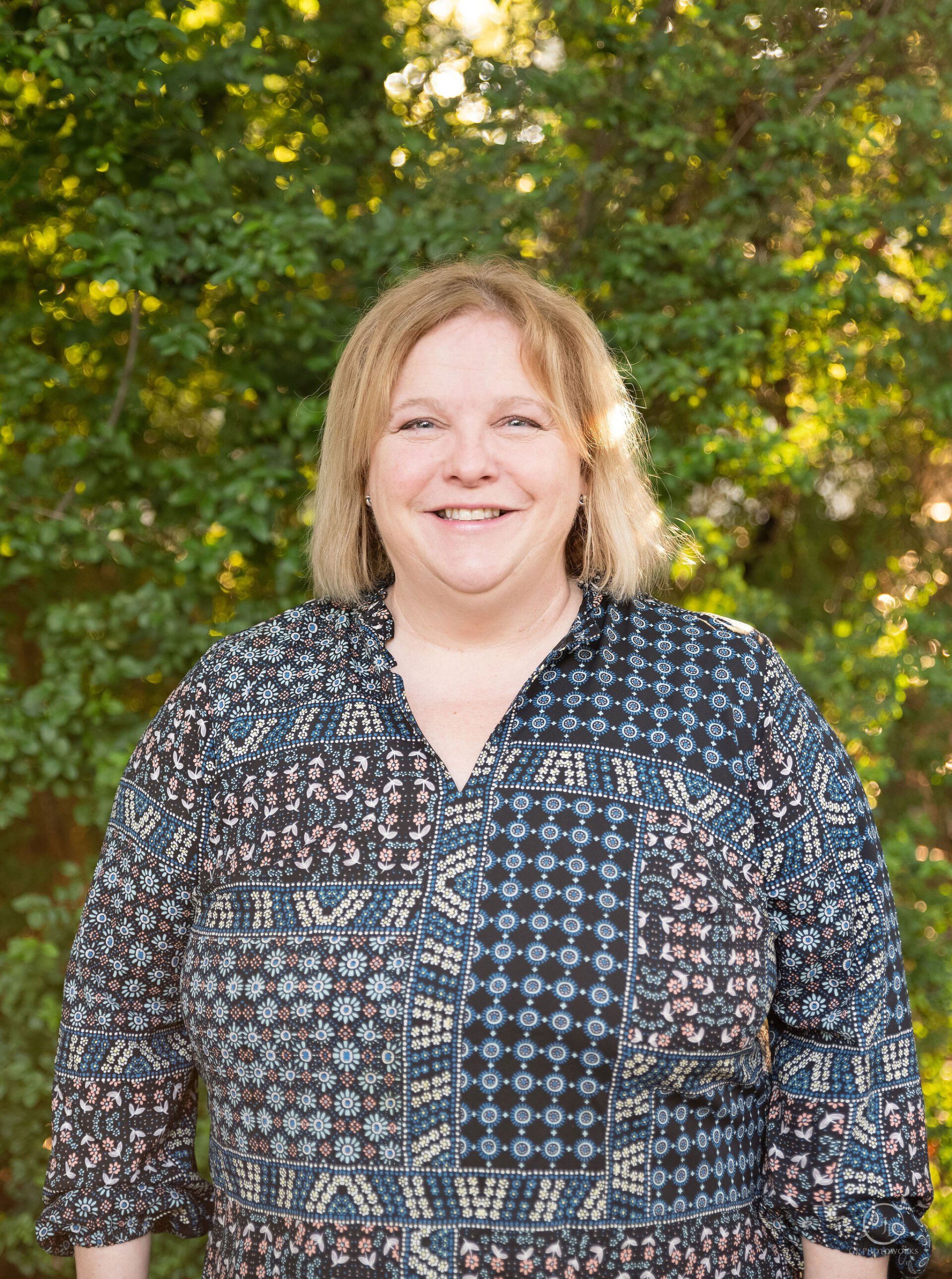 Smiling woman in a patterned blouse standing outdoors in front of green foliage