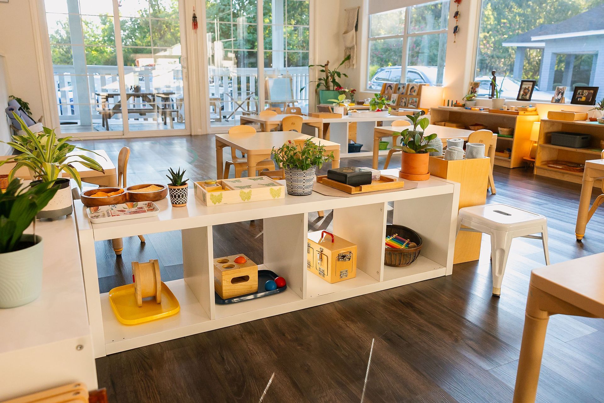 Bright classroom with plant-filled shelves, wooden tables, and large windows overlooking a sunny patio.
