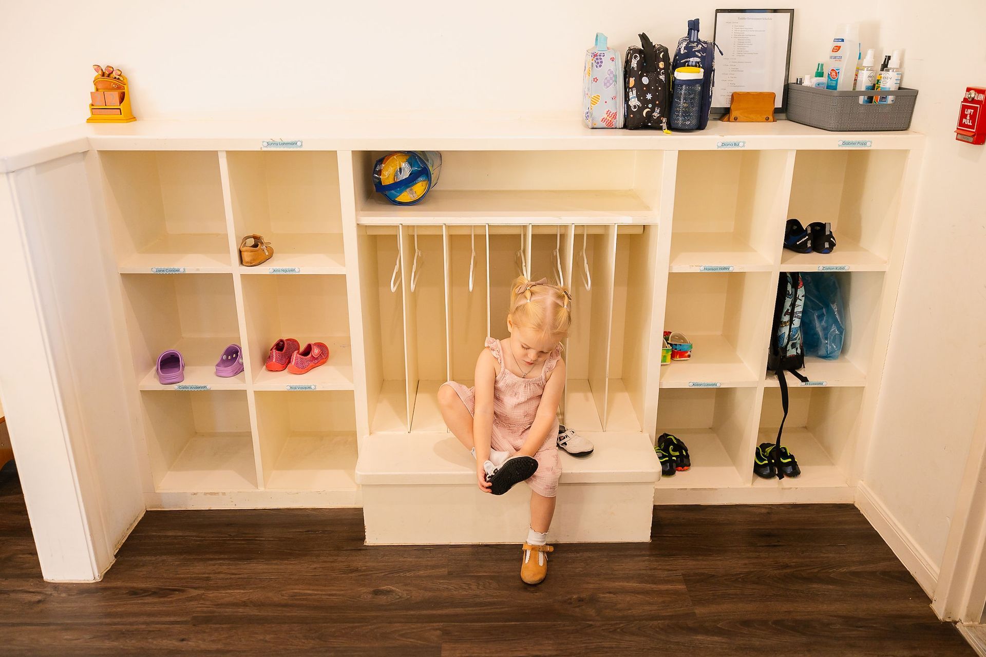 Child sitting on a bench in a cubby-lined locker room with shoes and bags on shelves
