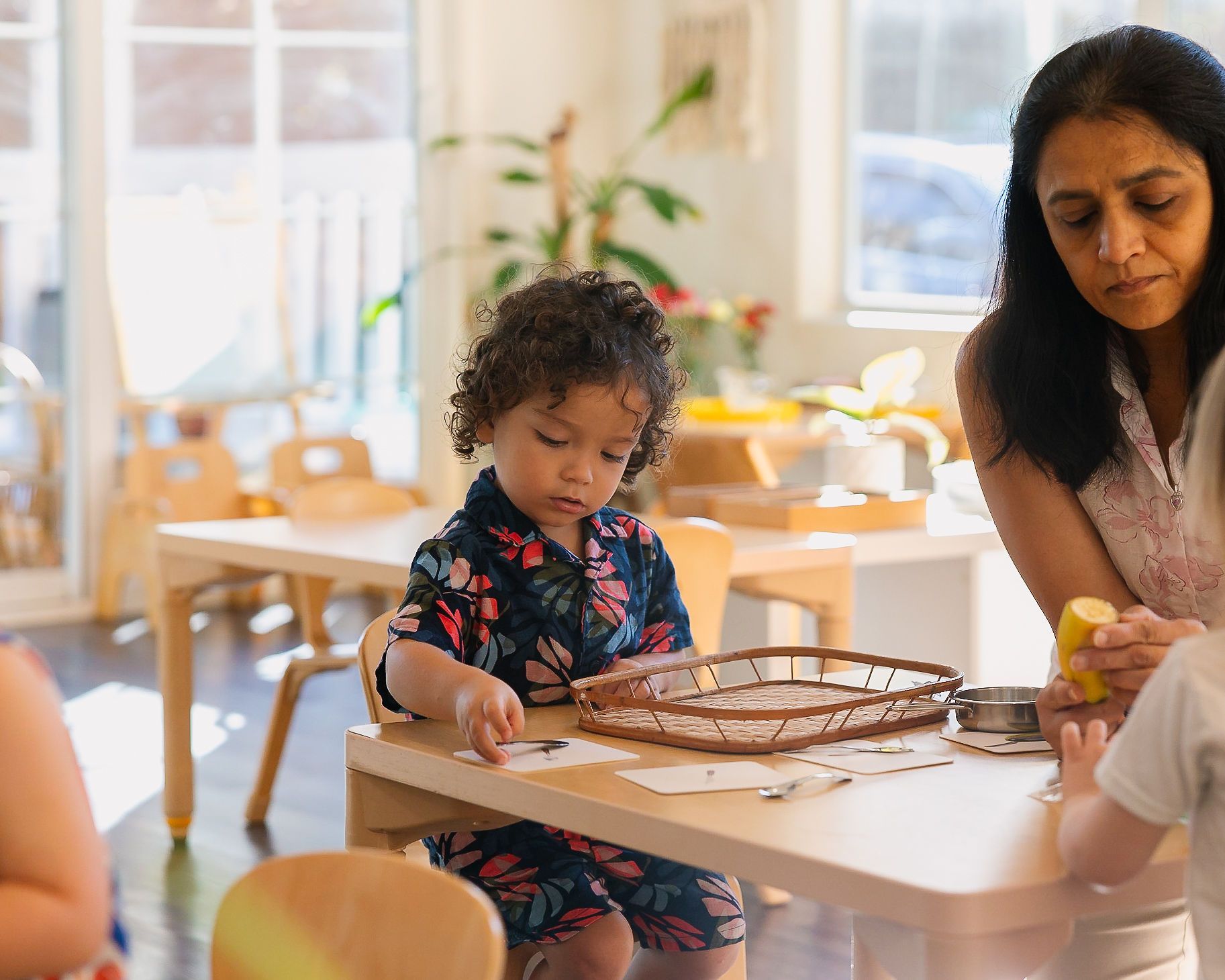 Child working with a tray at a table while an adult sits nearby in a bright room