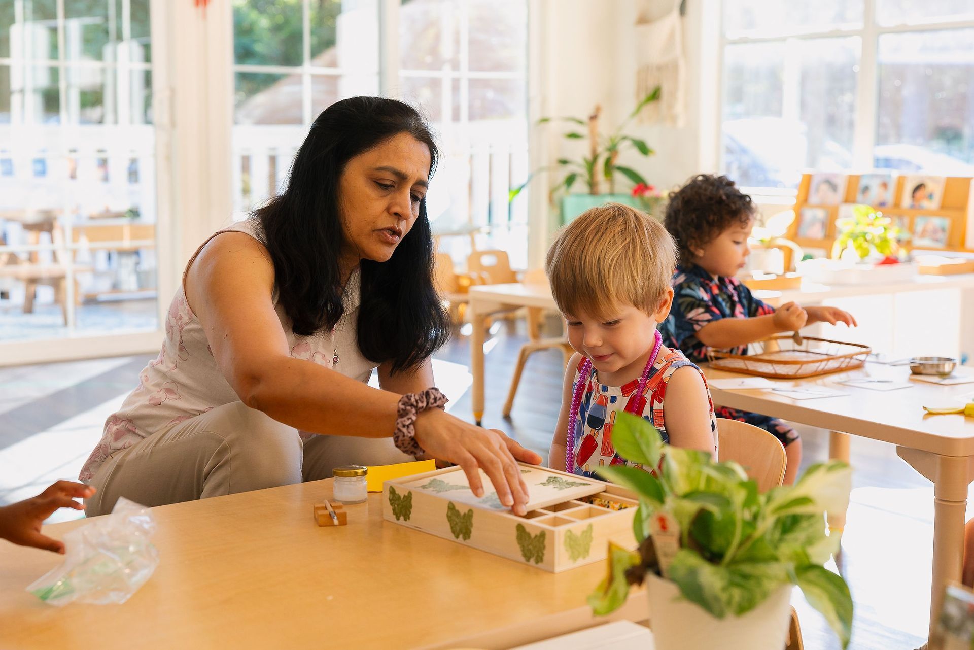 Adult helping child with a puzzle at a table in a bright classroom, with another child nearby.