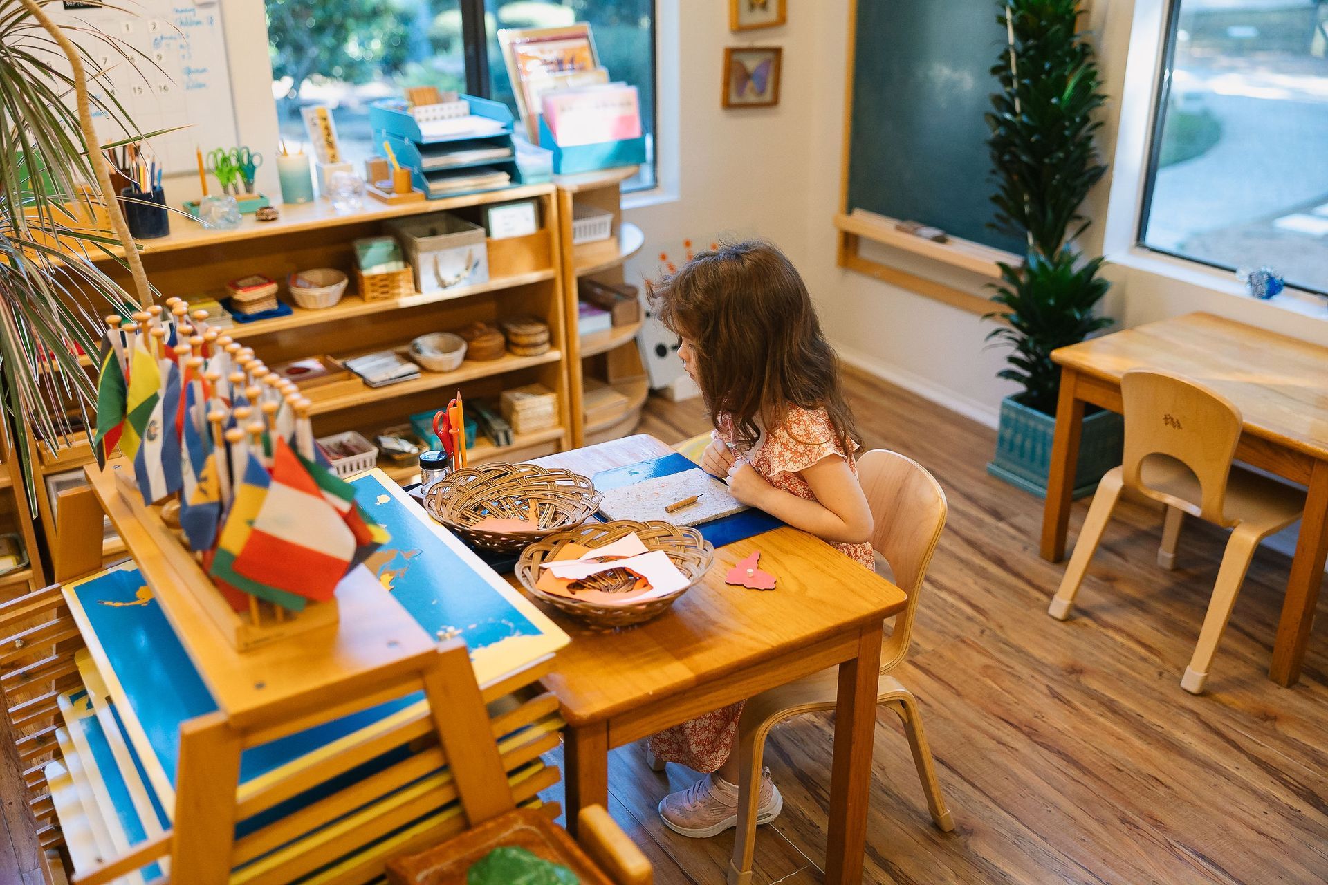 Child working at a small wooden table in a bright classroom with flags and art supplies.