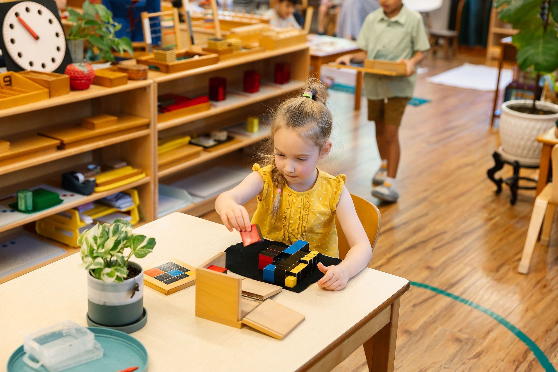 Child in a yellow dress working at a classroom table, with shelves and toys in the background