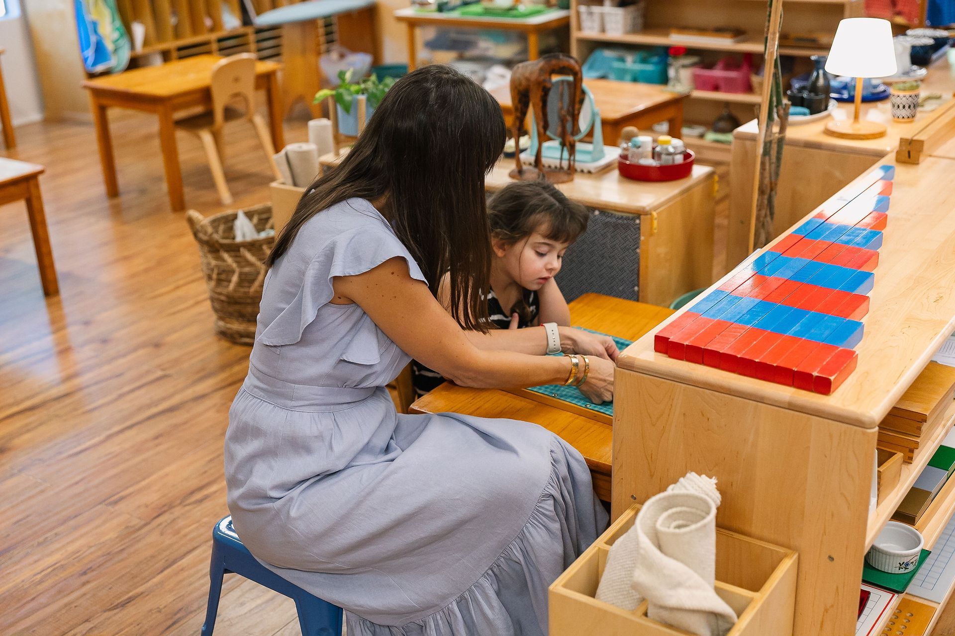 A guide showing a child how to use a dressing frame in a brightly lit Montessori classroom.