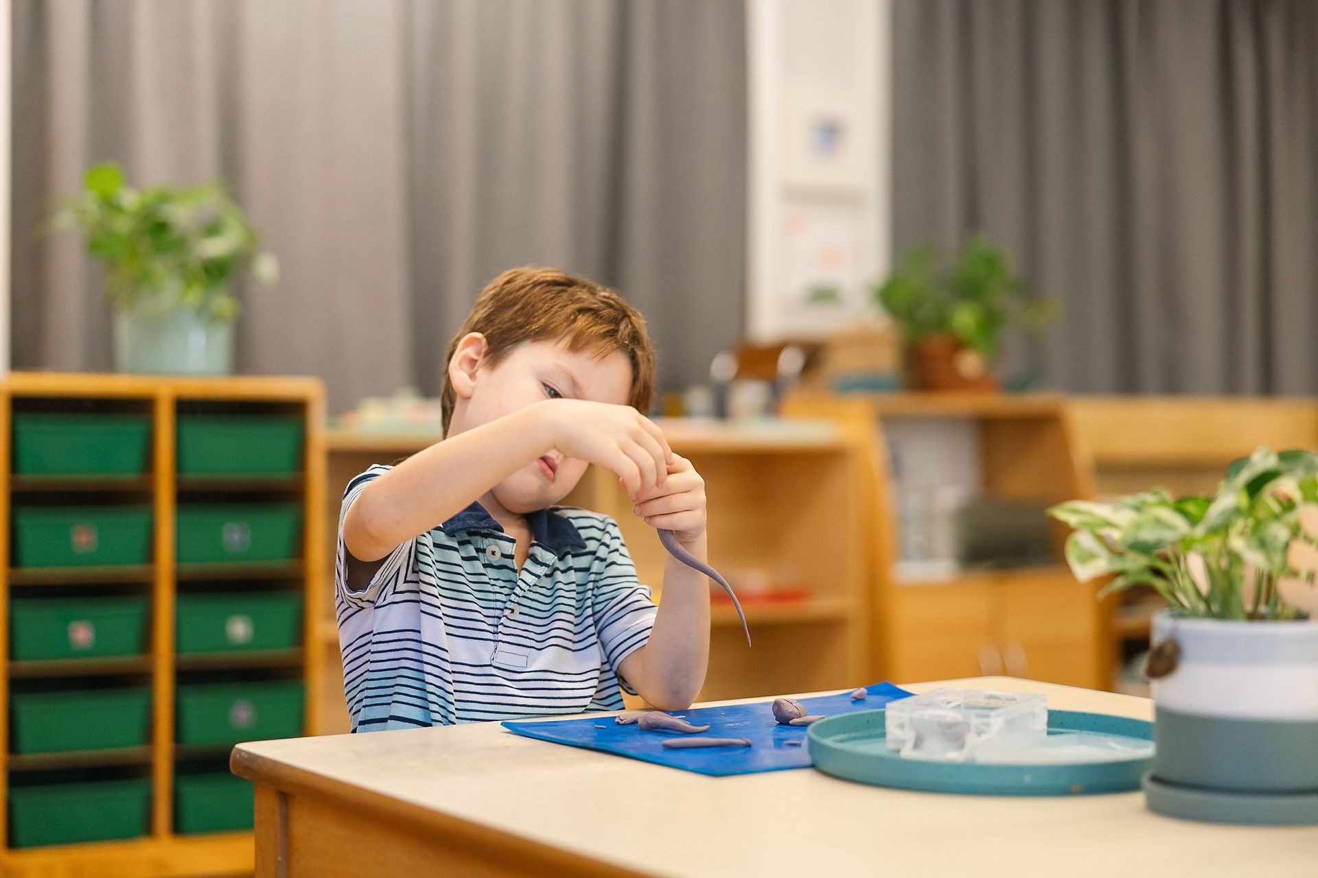 Child crafting with clay at a classroom table, with trays and potted plants in the background