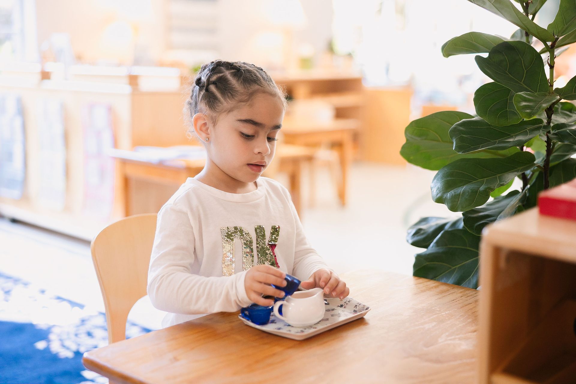 Child in a bright classroom pours into a small white cup at a wooden table.