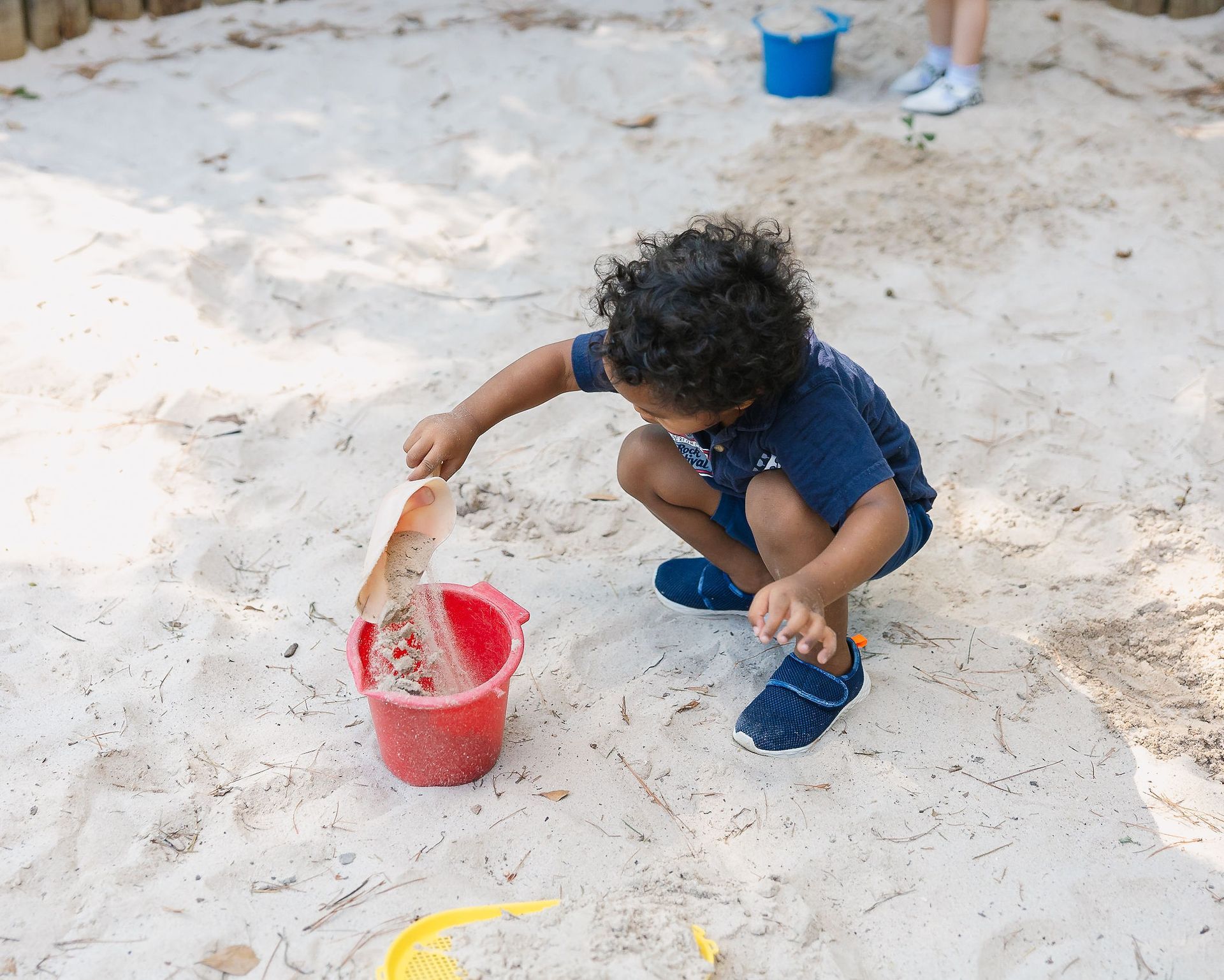 Child scooping sand into a red bucket in a sandbox with blue toys nearby