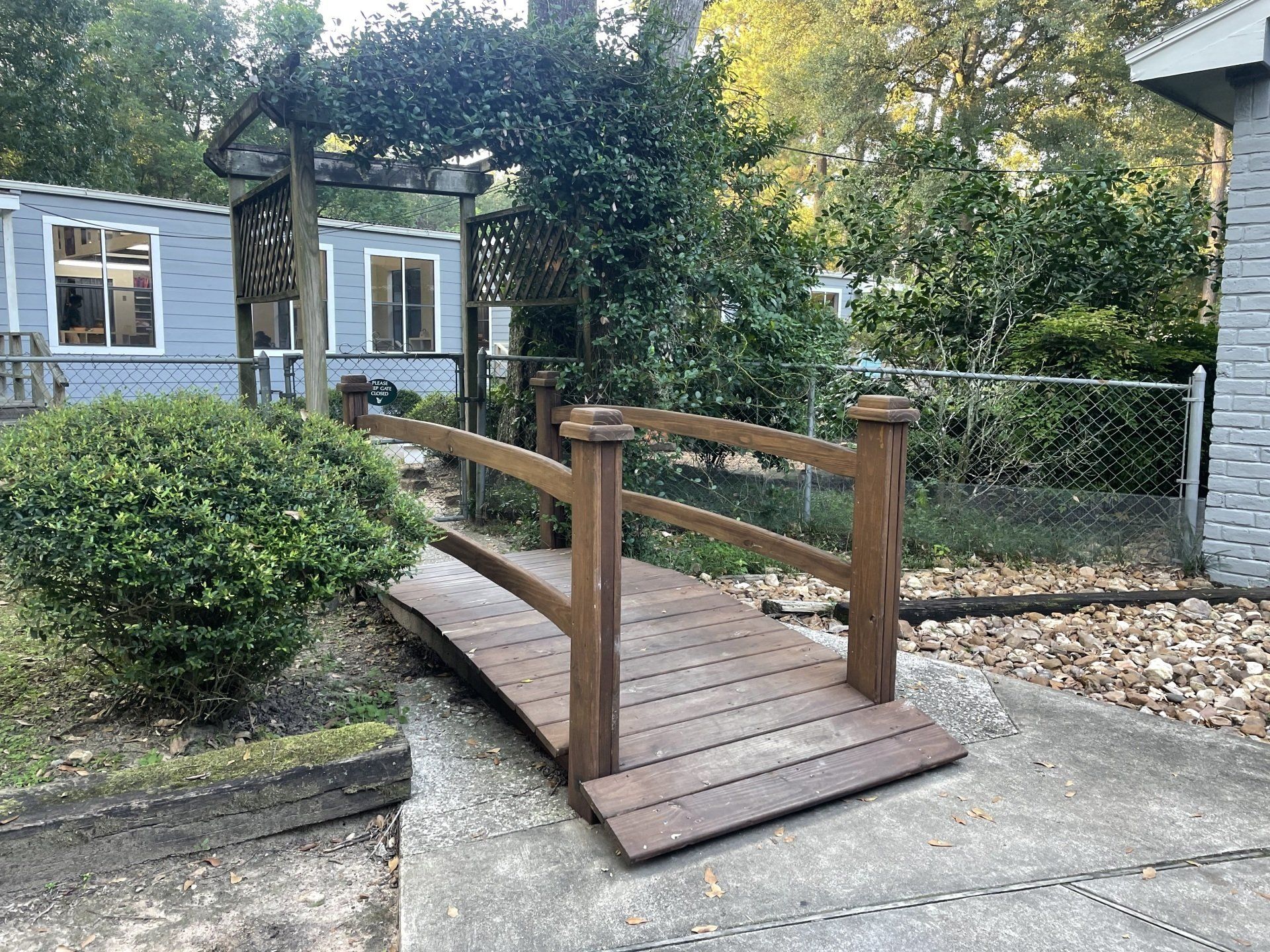 At Northwood Montessori School a wooden bridge over a small incline, leading toward a blue building with a trellis.