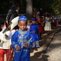 Montessori Children in costumes, holding flags, march outside. Blue and white attire prominent.