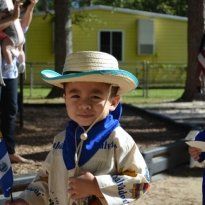 Montessori Boy in straw hat and traditional clothing, holding a flag, smiling outside a yellow building.