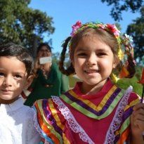 Montessori Girl in colorful dress and flower crown smiles, boy beside her, woman in background.
