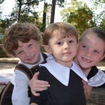 Three Montessori children smiling, embracing outdoors. One child wears a costume with a white collar.