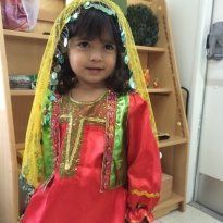 Young Montessori girl in red dress and yellow head covering smiles in front of shelves.