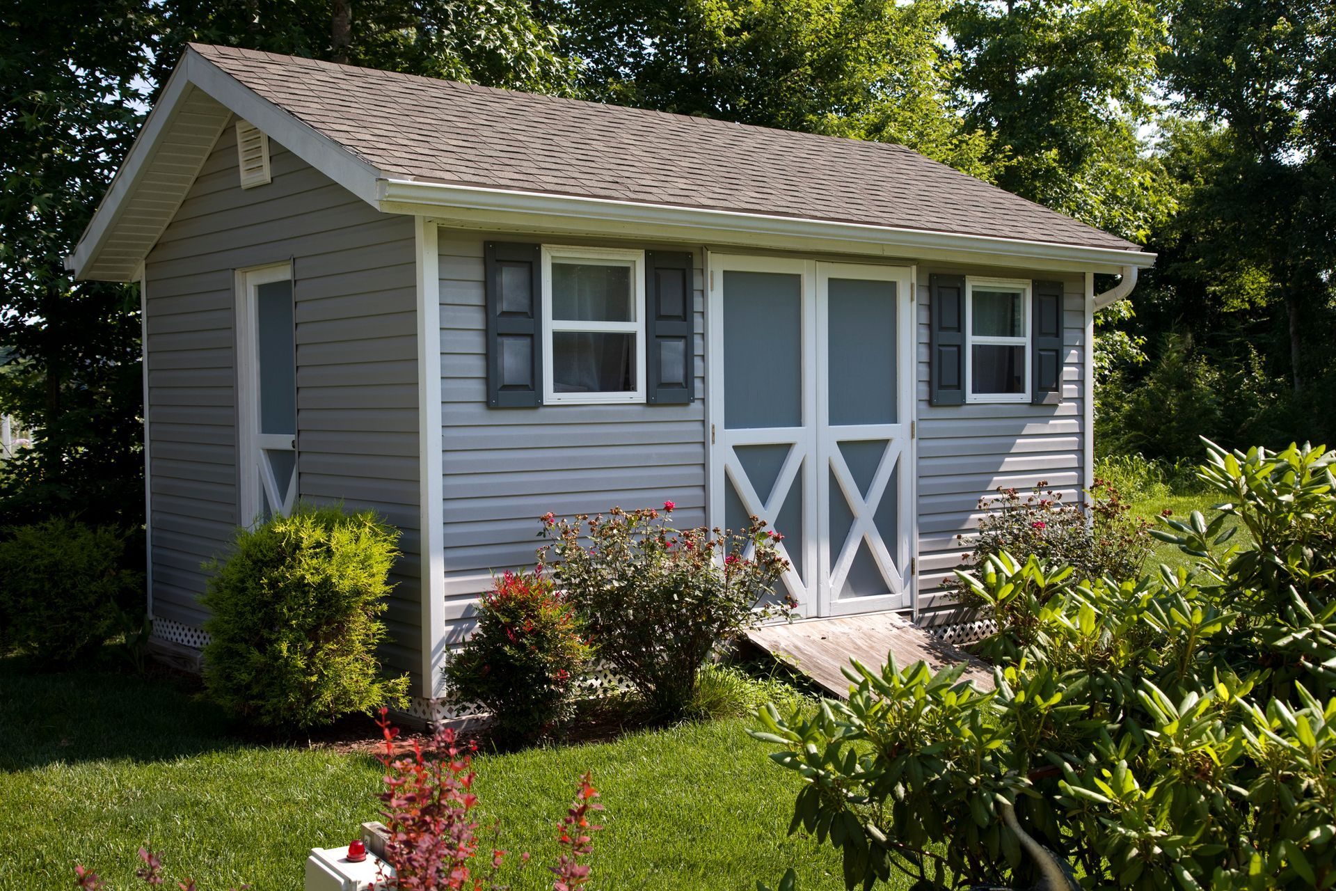Small gray shed with white trim, double doors, two windows, and dark shutters; surrounded by bushes and grass.