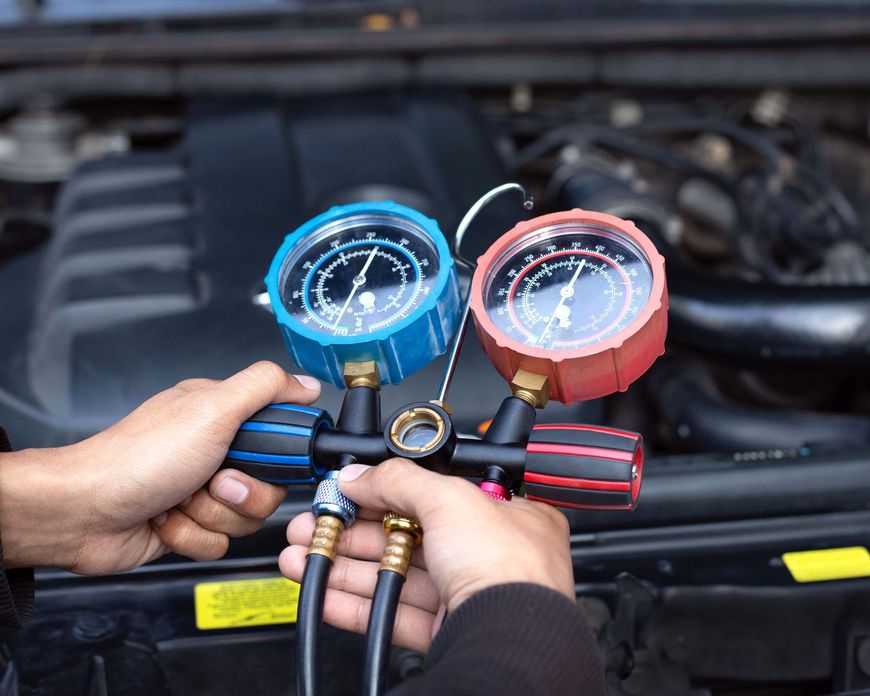 Mani che reggono manometri blu e rossi collegati ai tubi dell'aria condizionata dell'auto sotto il cofano