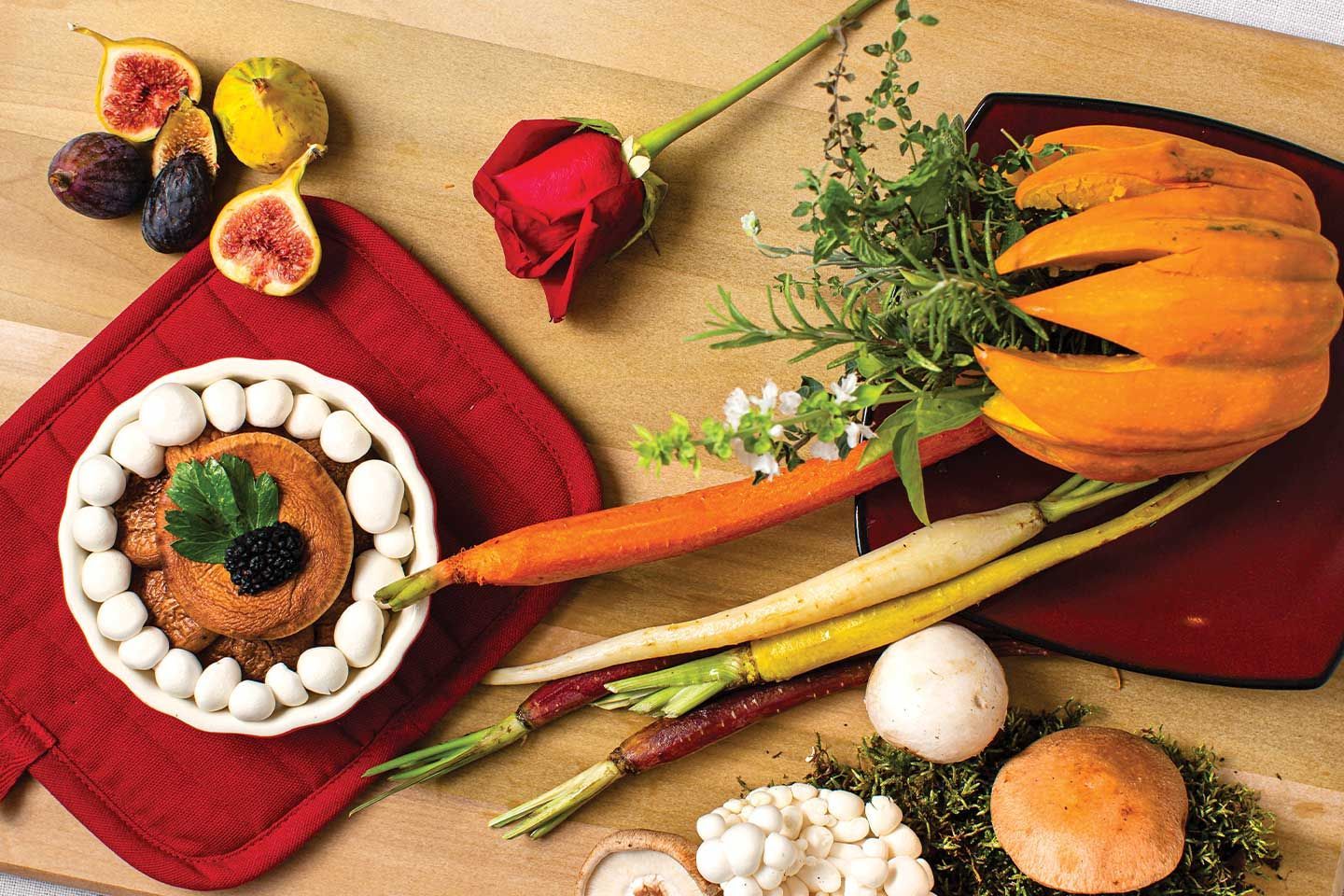 Still life: Food arrangement with squash, carrots, figs, mushrooms, herbs, and a rose on a wooden surface.