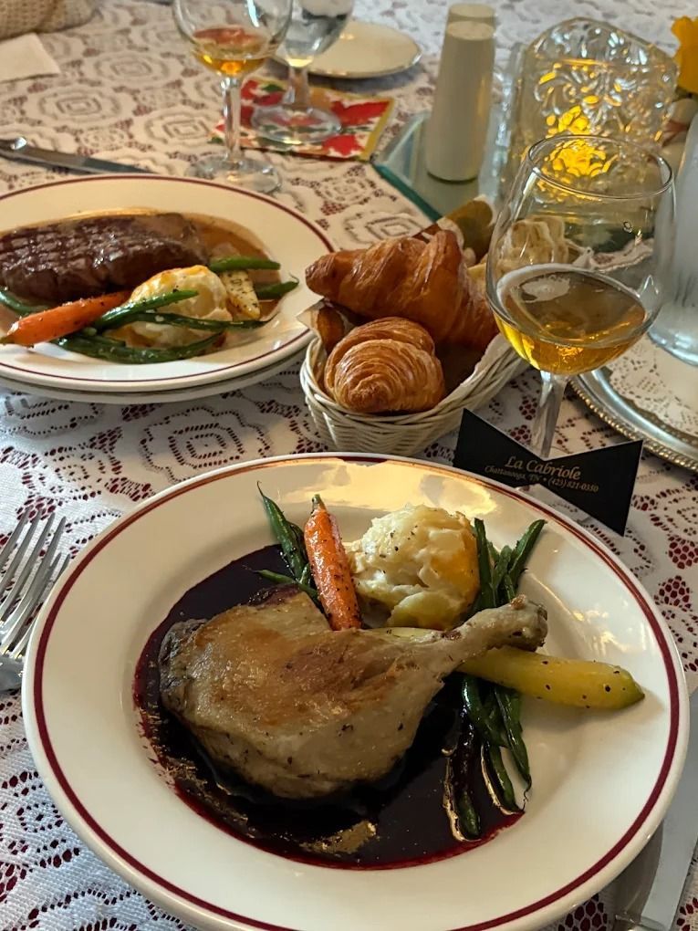 Two plates of food, duck confit and steak, with croissants and wine on a table set for dining.