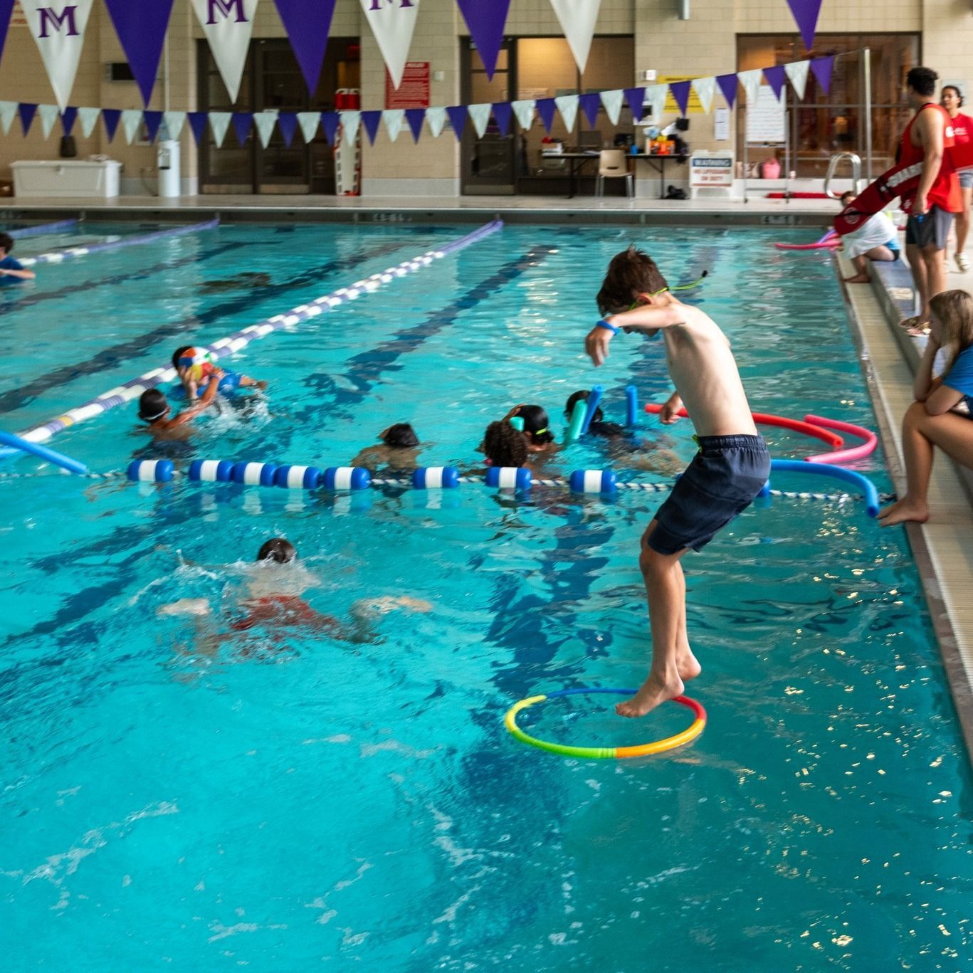 A boy jumps into a swimming pool with a purple flag that says m on it