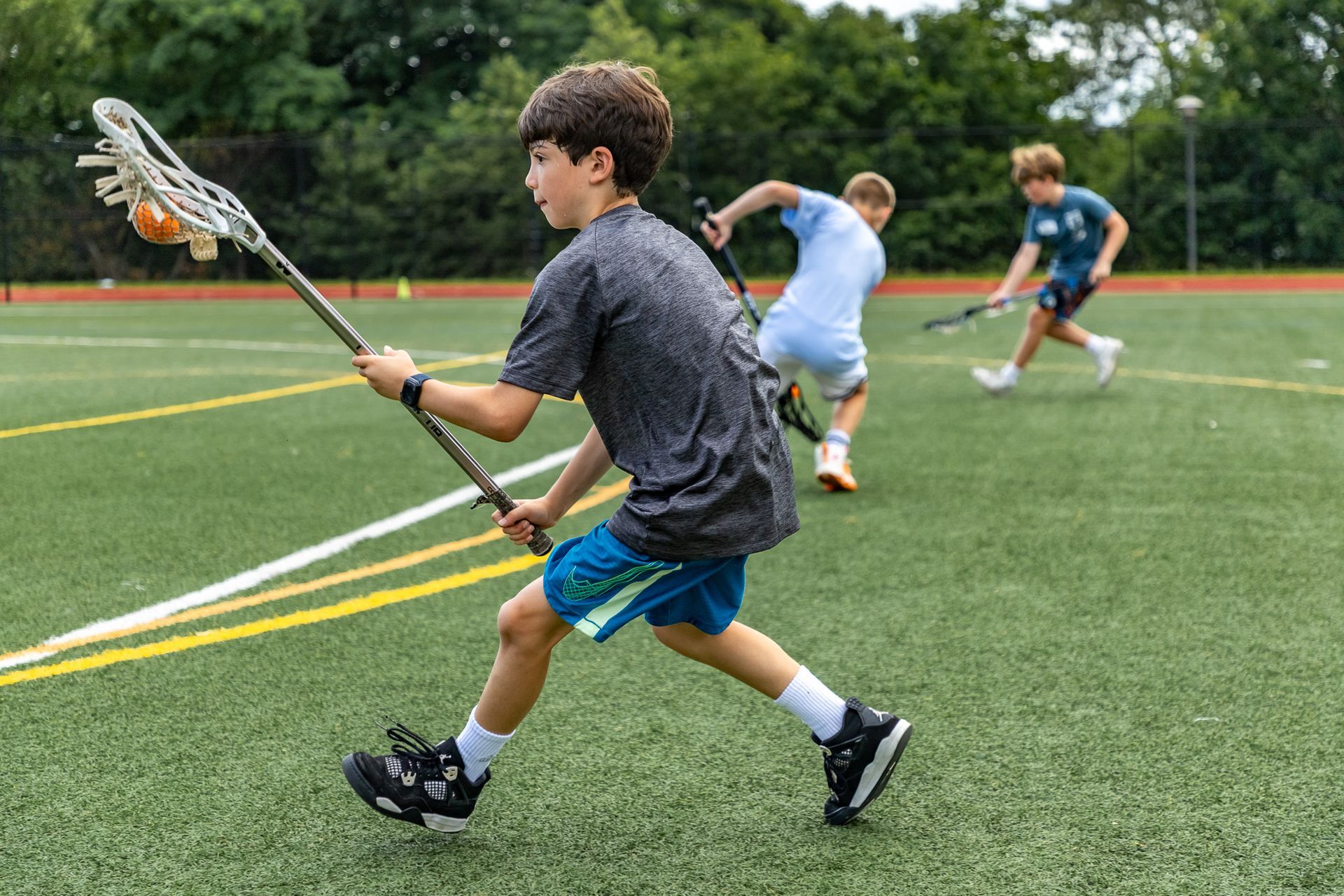 Boy in gray shirt and blue shorts playing lacrosse on a green field, holding a lacrosse stick with an orange ball.