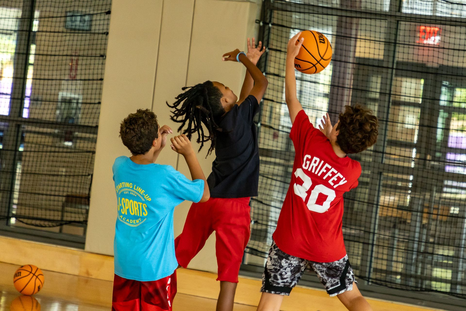 Three kids playing basketball indoors; one jumps for the ball, while the other two reach up to try to grab it.