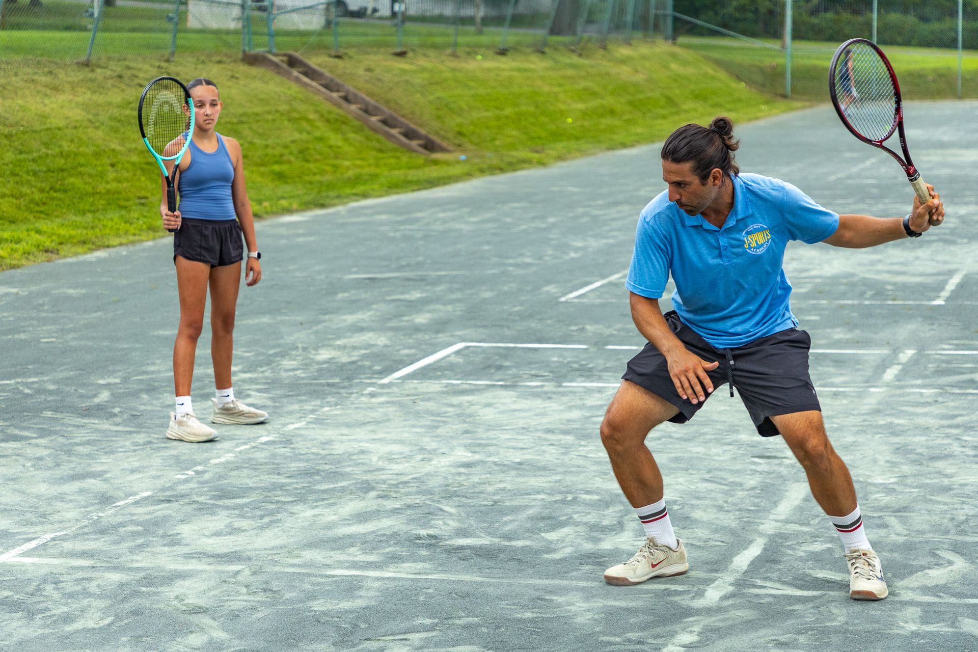 Woman and man playing tennis on a gray court; man swings racket, woman ready.