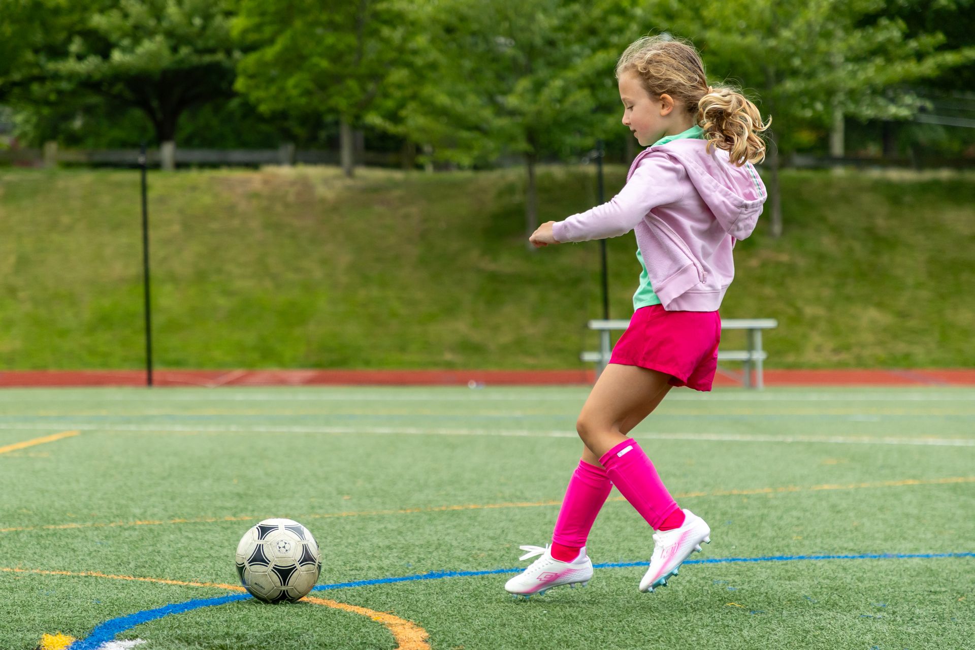 Girl in pink kicking a soccer ball on a green field.