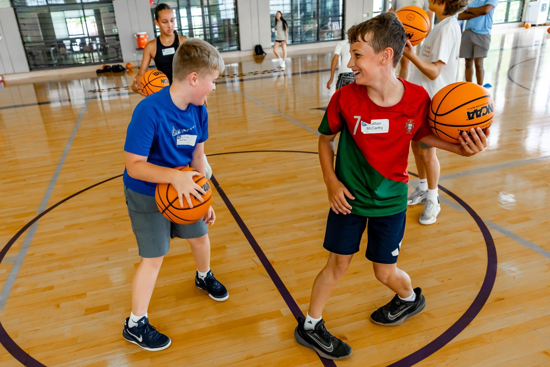Two young boys with basketballs on a court, smiling, one in blue, the other red and green. Others play nearby.