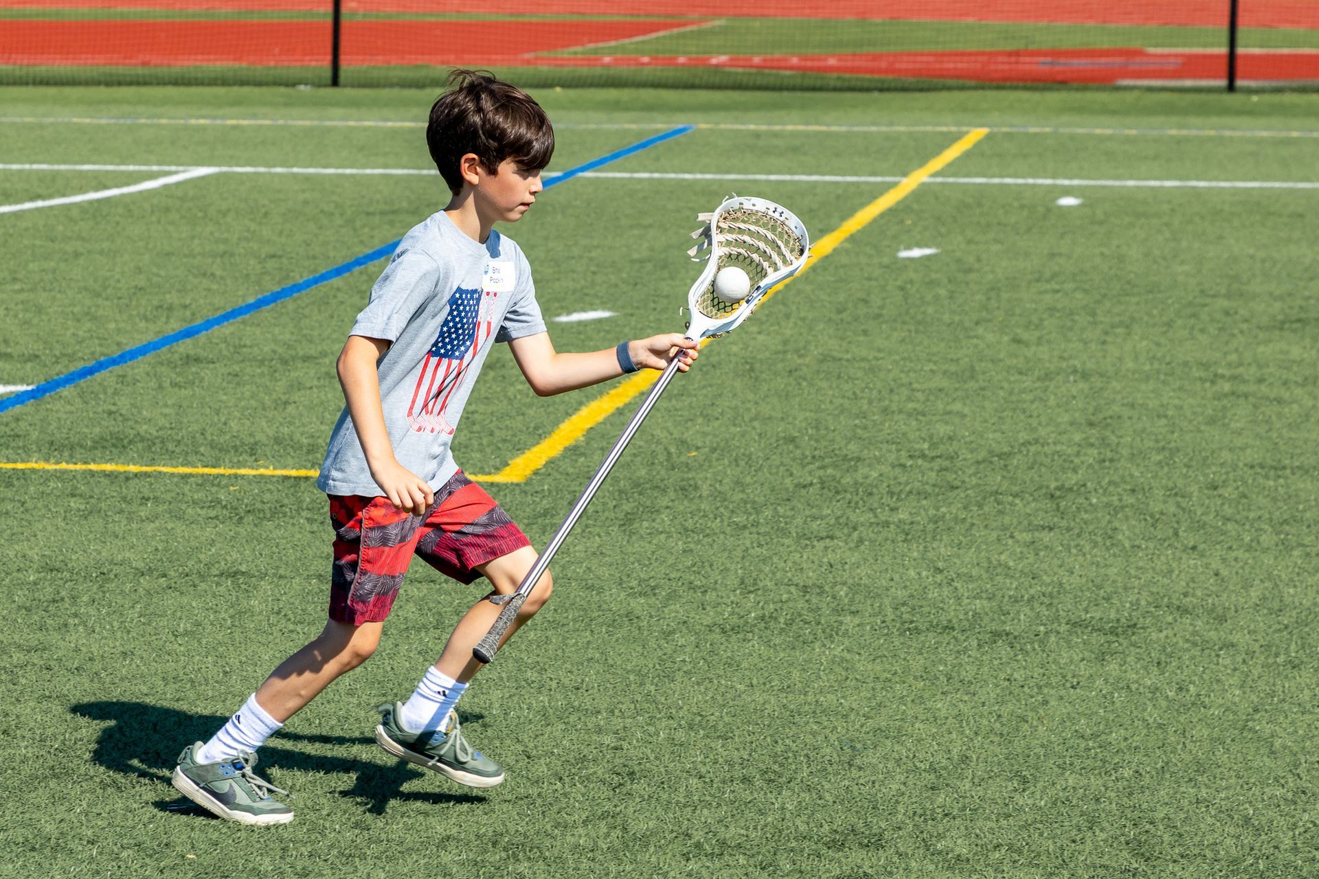 Boy in red shorts and American flag shirt runs with lacrosse stick on a green field.