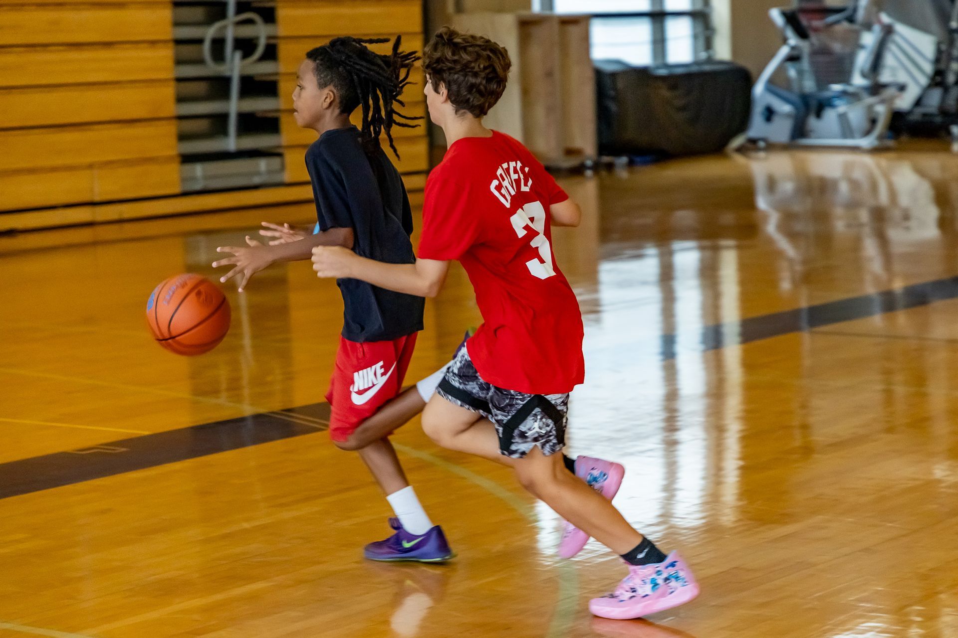 Two young basketball players dribble on a wooden court; one in red, one in black.