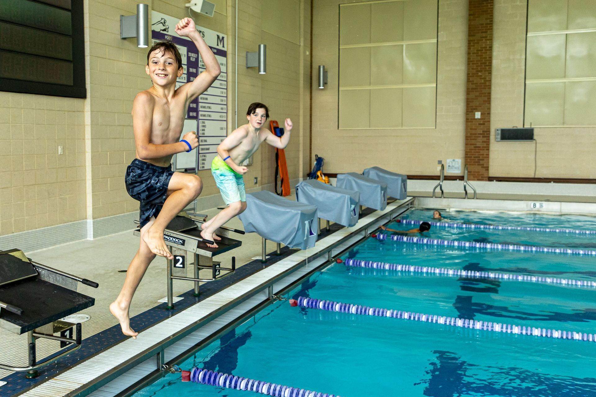 Two boys jumping excitedly into a swimming pool. Indoor pool, with blue water.