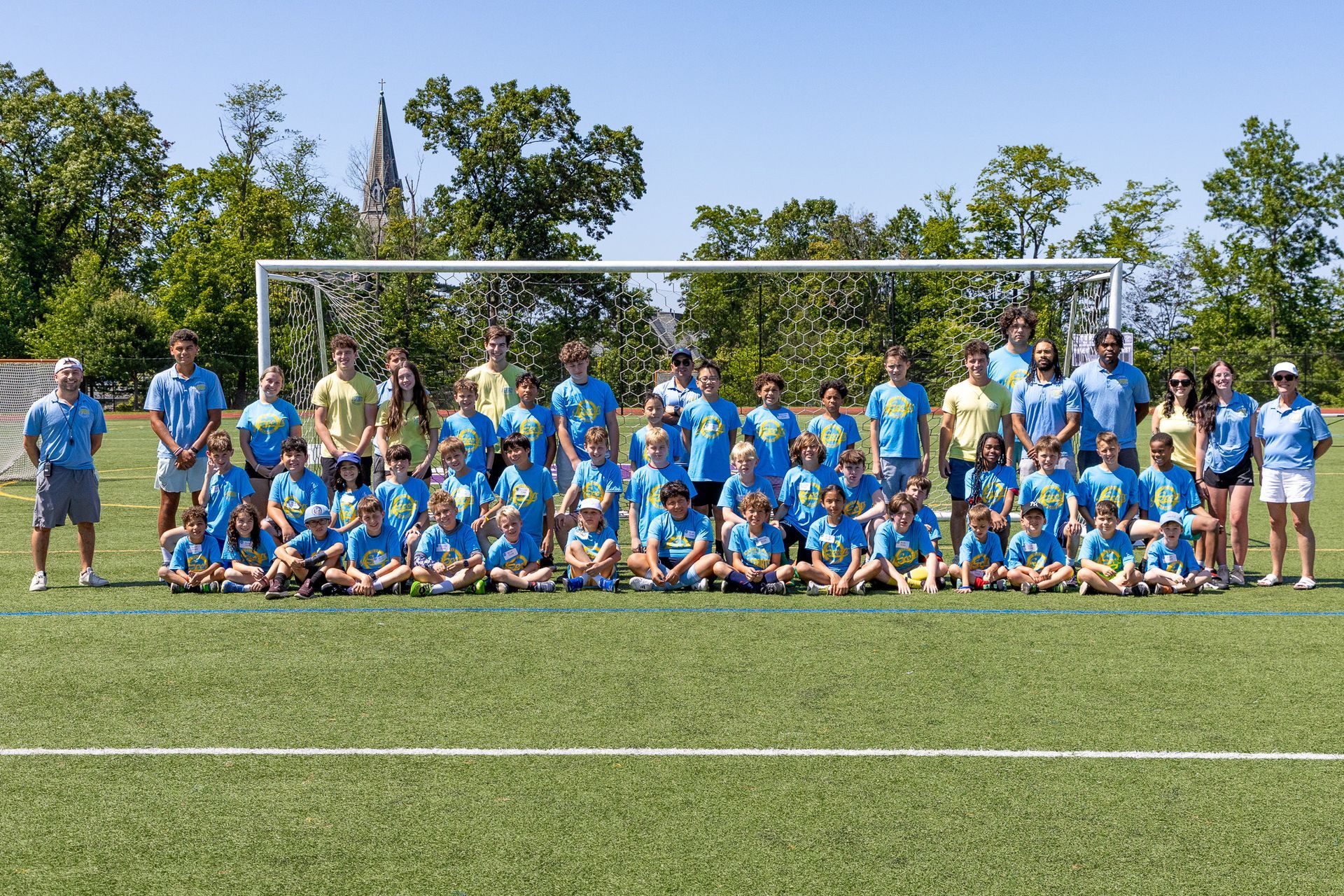 Large group poses on a soccer field, wearing matching blue and yellow shirts, in front of a goal. Sunny day.