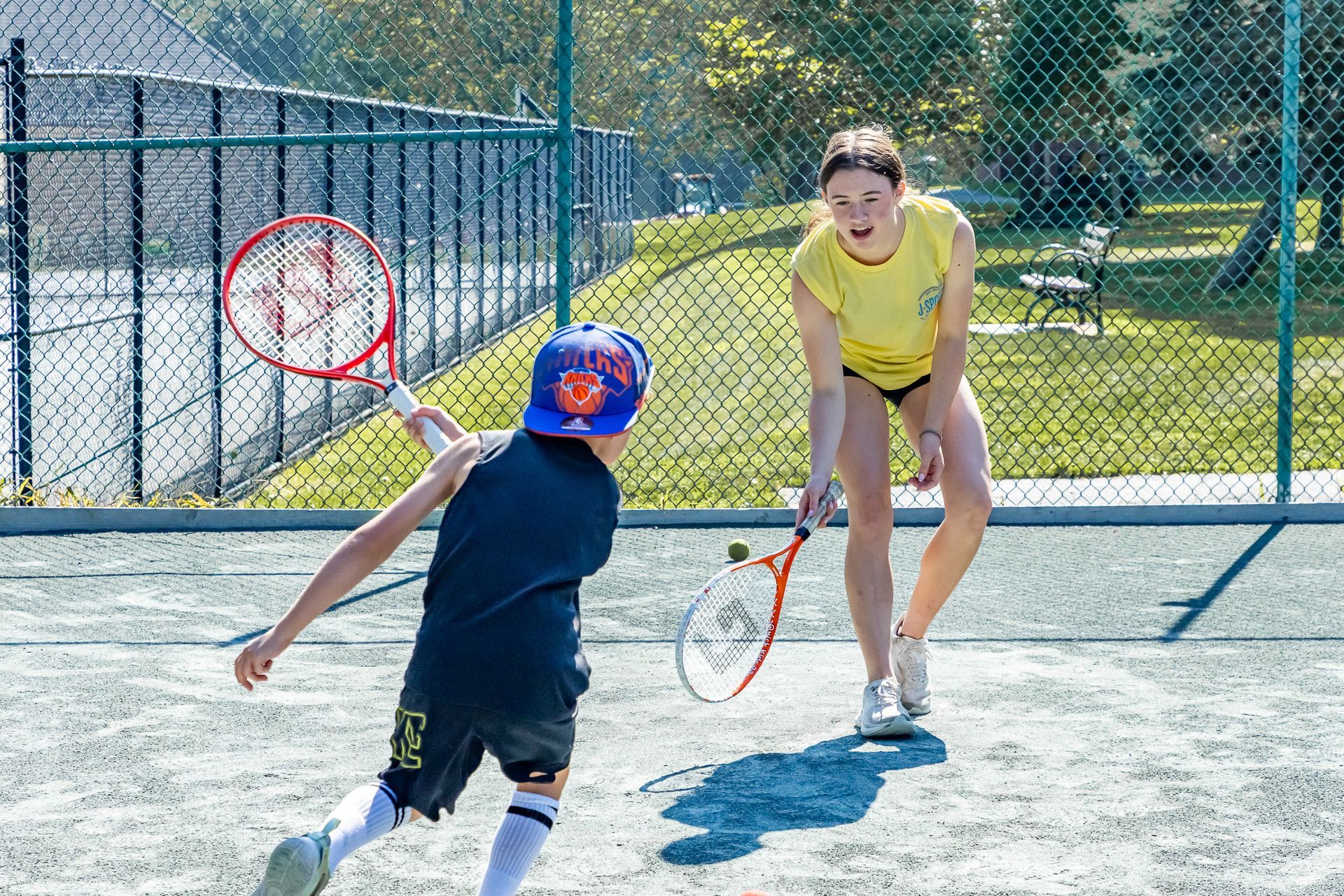 Child playing tennis with a coach on an outdoor court; child swings racket, coach watches.