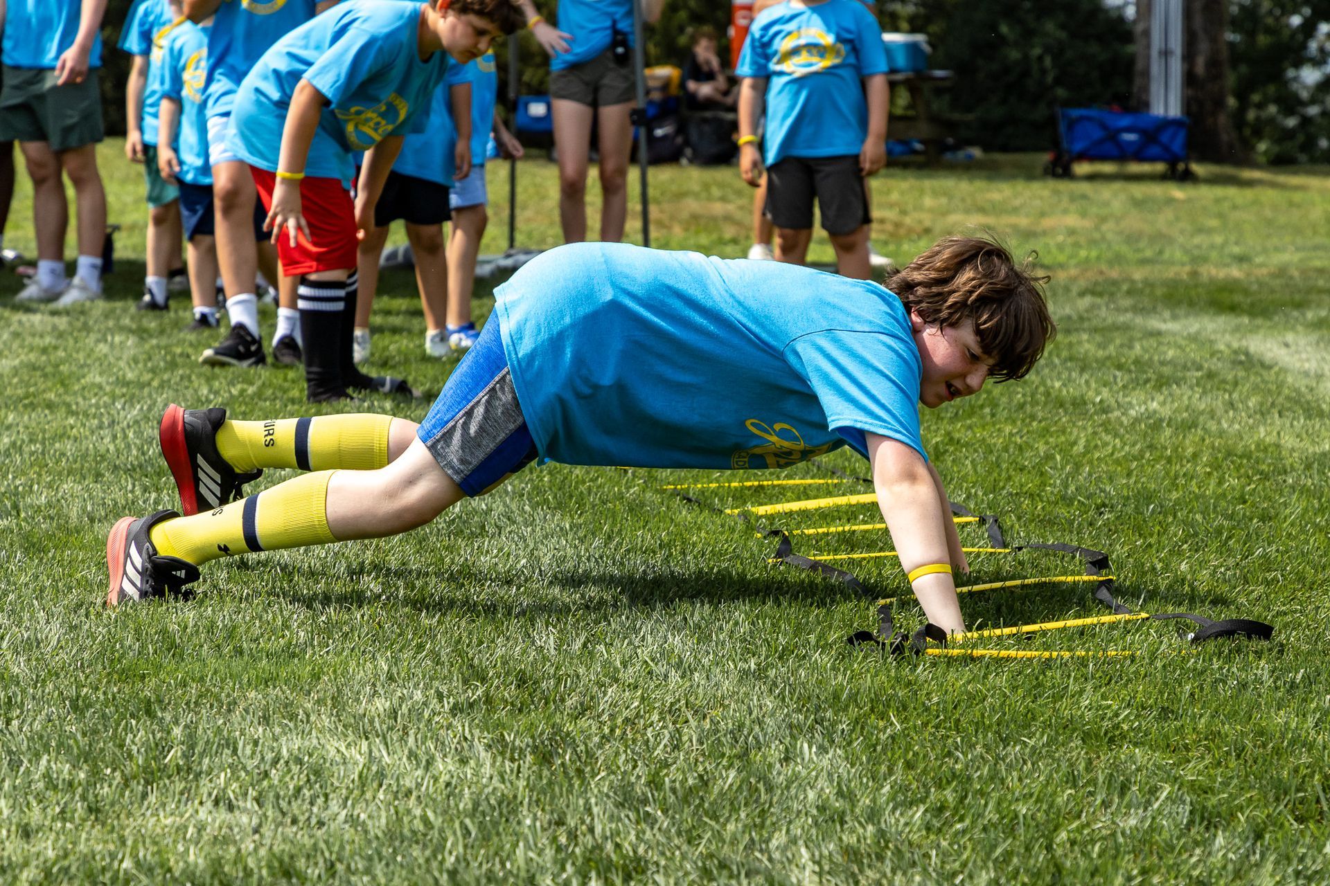 Boy in blue shirt doing push-ups on a training agility ladder on grass, other children in background.