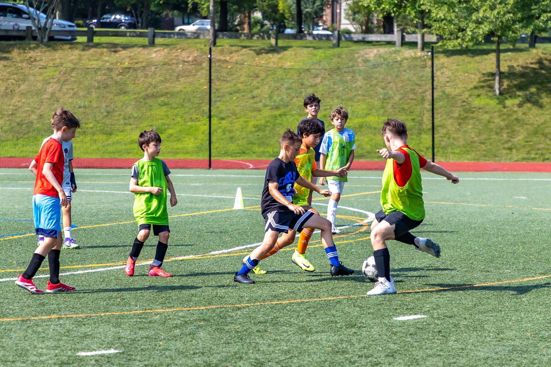 Kids playing soccer on a green field; some wear red, green, and blue jerseys. Sunny day.
