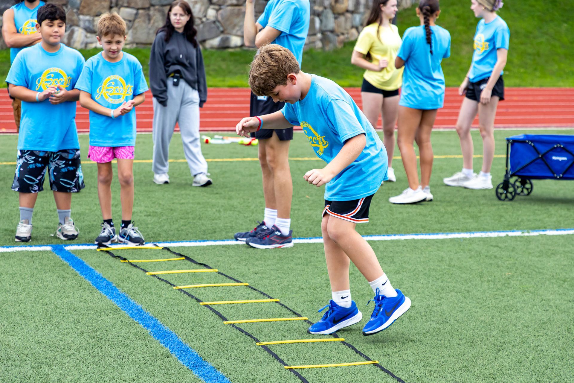Children in blue shirts doing agility ladder drills on a track.