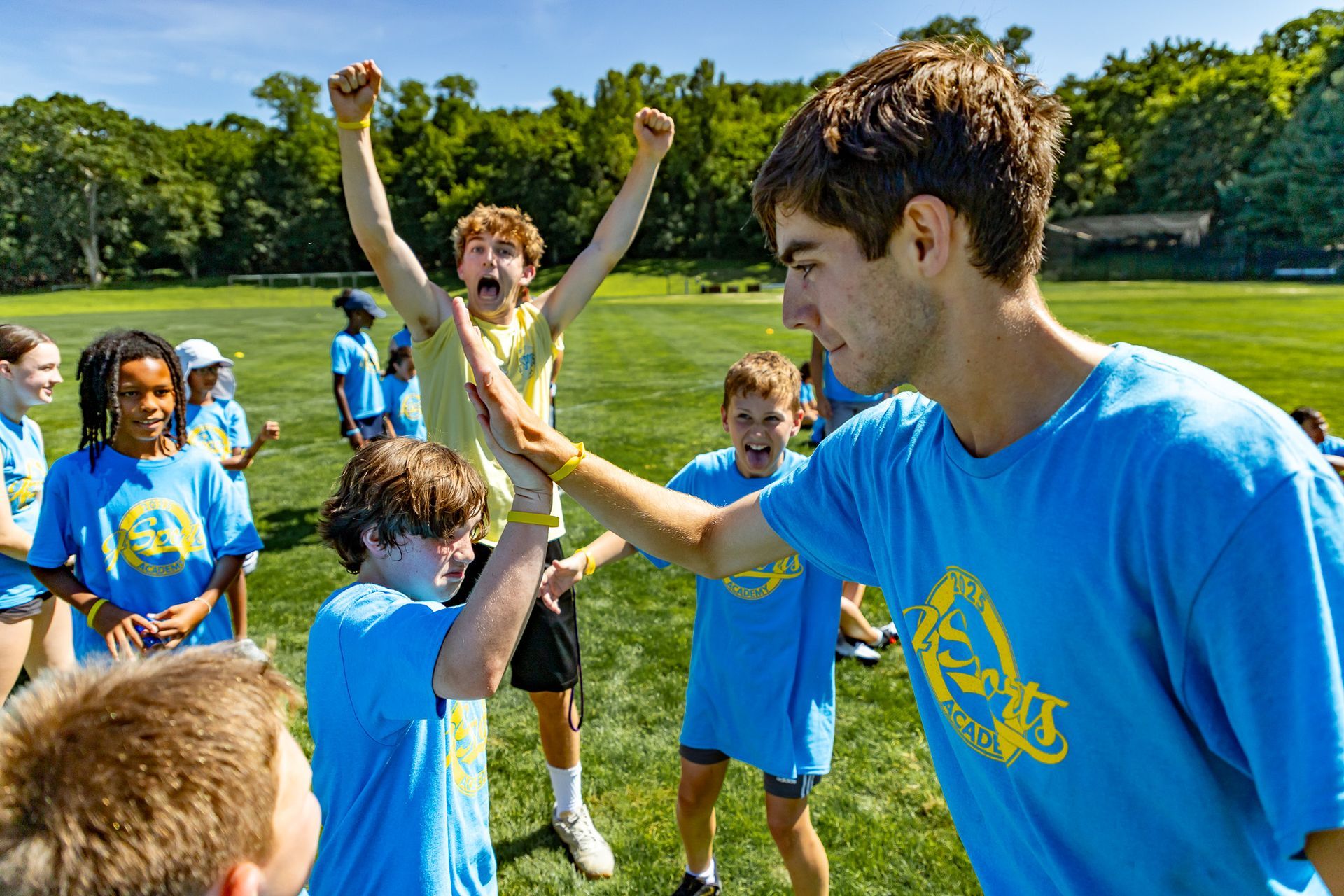 Group of children in blue shirts celebrating on a green field, high-fiving. One boy raises arms in excitement.