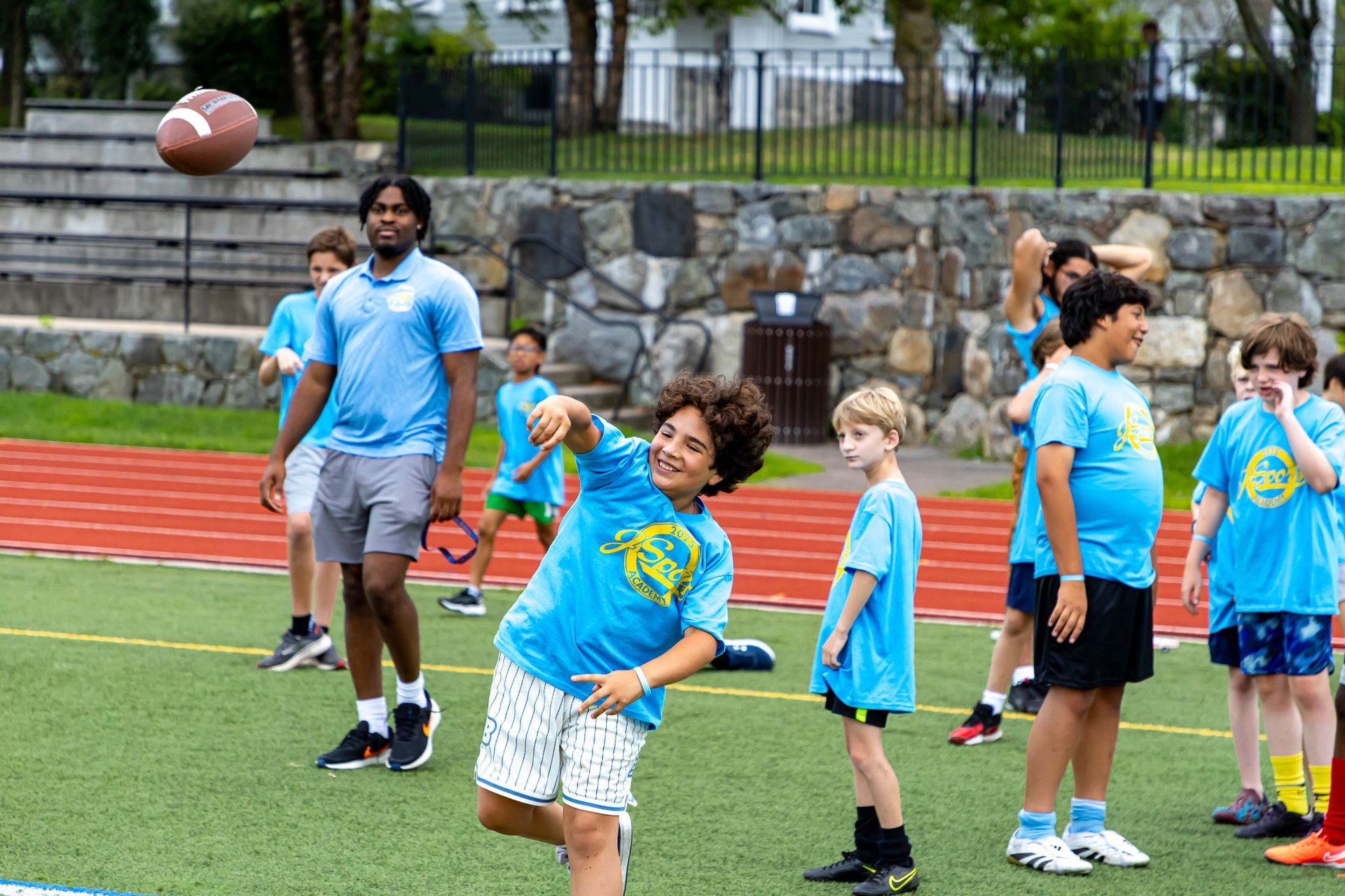 Boys throwing a football during an outdoor activity. They're wearing blue shirts. A coach watches.