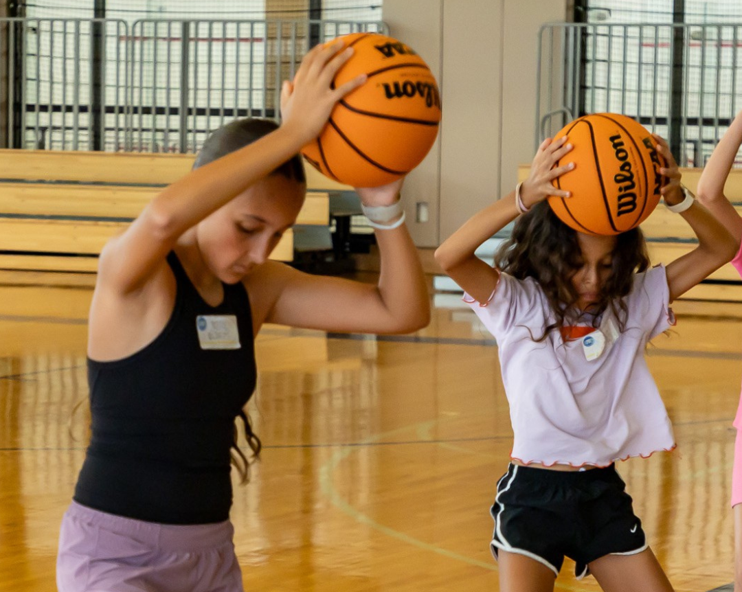 Two girls in a gym balancing basketballs on their heads, arms bent, focused expressions.