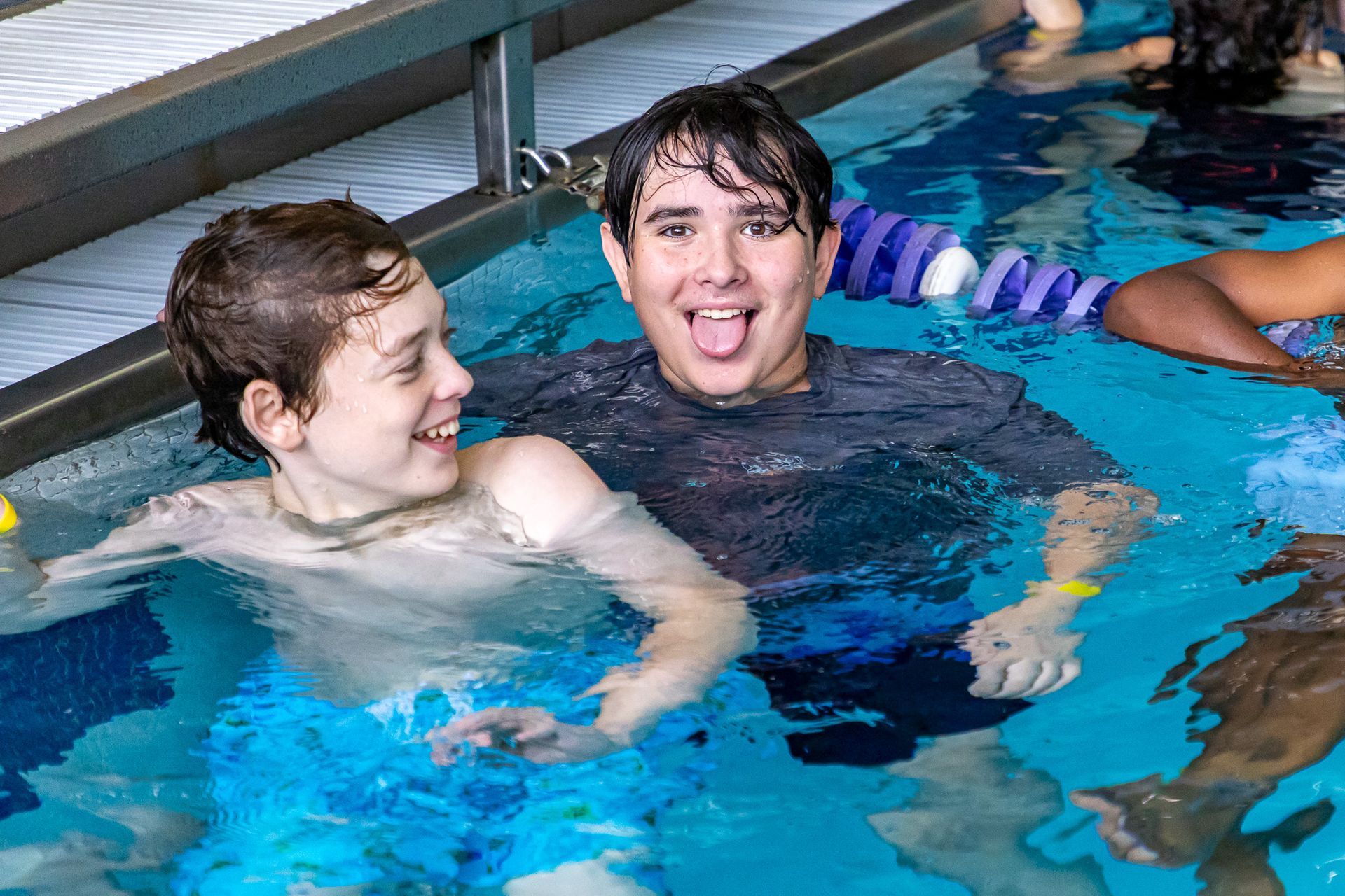 Two boys in a pool, smiling. One sticks out tongue. Blue water, indoor setting.