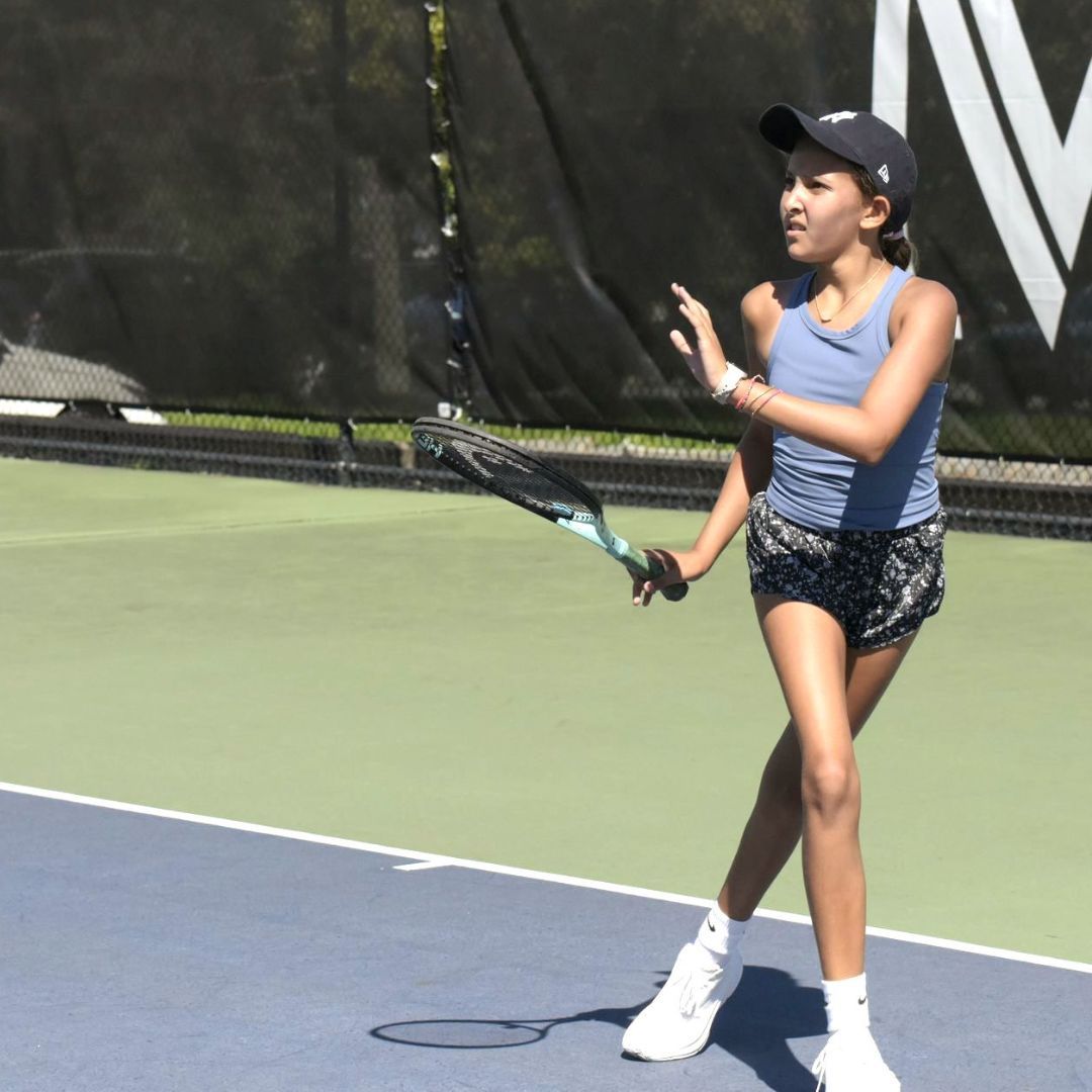 A young female tennis player wearing a blue tank top and shorts swings a racket on a court.