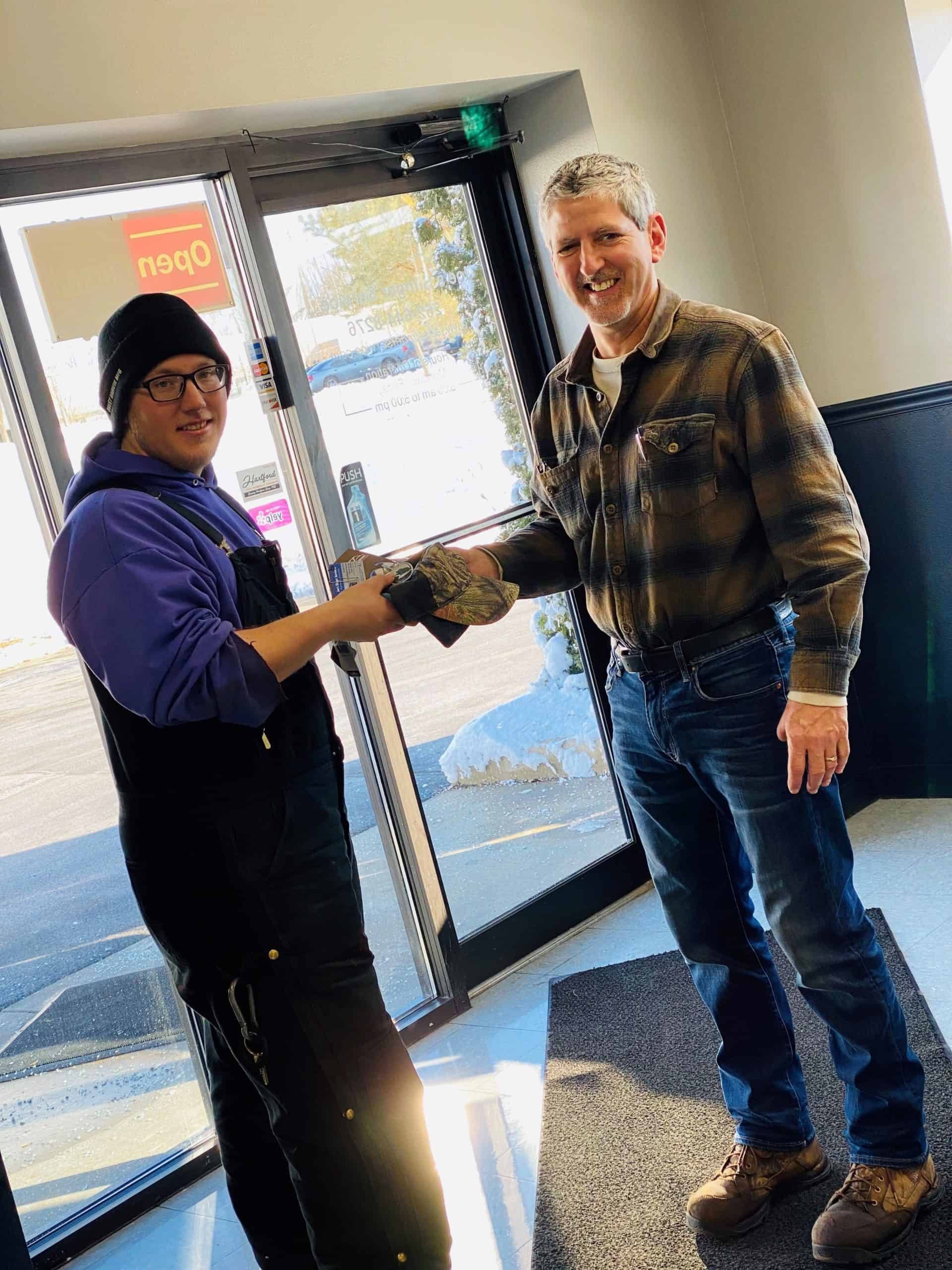 Two men shaking hands in a doorway, one wearing overalls and a beanie, the other a plaid shirt.