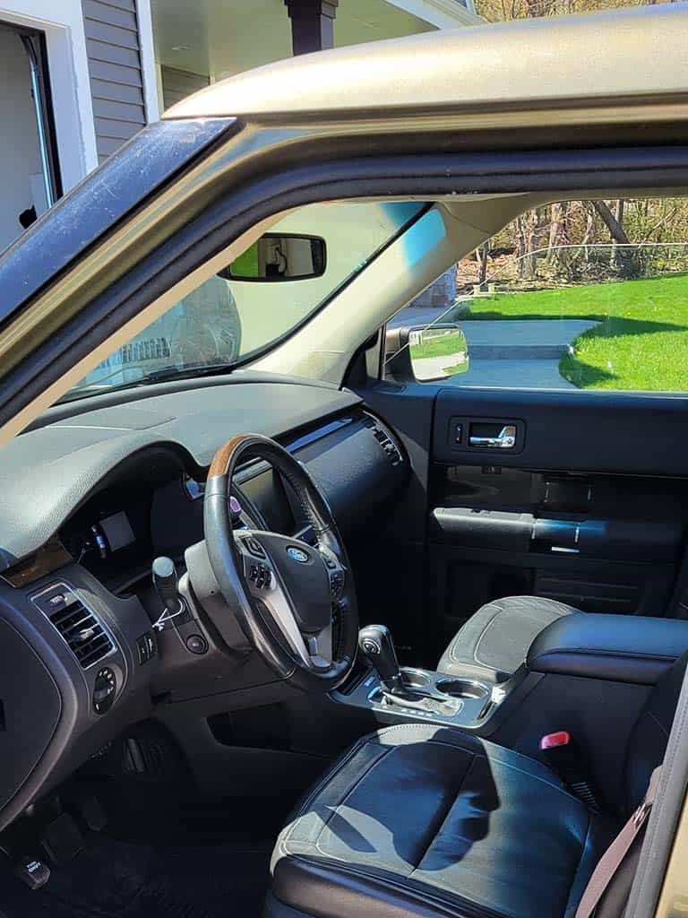 Interior view of a beige Ford Flex, showing the dashboard, steering wheel, seats, and outside view through the open door.