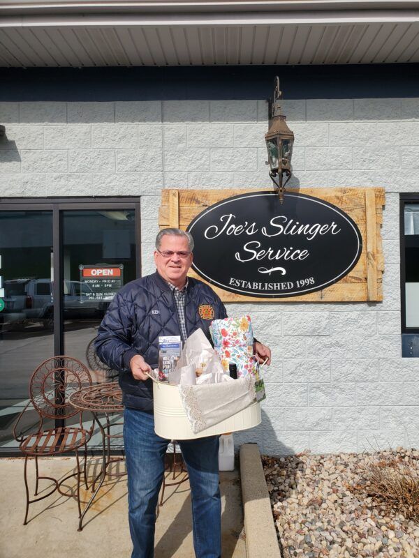 Man holding a gift basket stands outside Joe's Slinger Service sign. He is wearing a blue jacket and jeans.