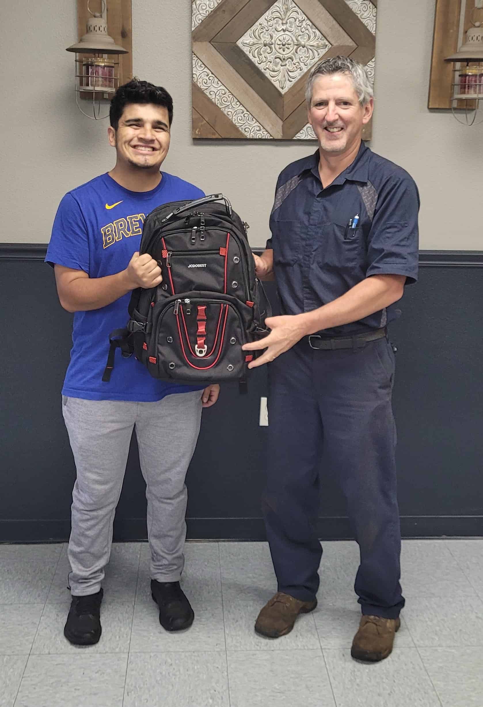 Two people holding a backpack, one smiling. Indoors, neutral background.