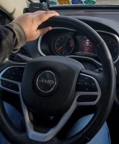 Person's hand on a Jeep steering wheel, dashboard visible. Black steering wheel, Jeep logo, inside a vehicle.