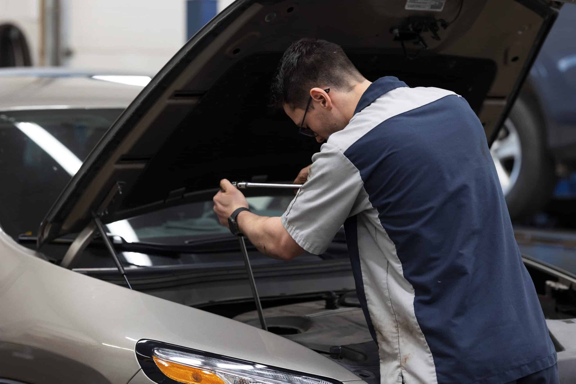 Mechanic working on car engine in a garage; gray car, wearing blue and grey uniform.