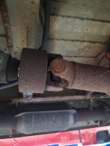 Underside view of a rusty vehicle's driveshaft, showing U-joint and connection to a large coupling.